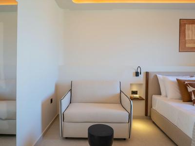 Bright hotel room with white armchair, bed, bedside table, and modern lighting.