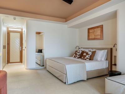 Bright hotel room with large bed, patterned pillows, and simple modern furniture.