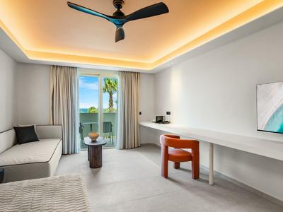 Bright hotel room with a couch, large table, chair, and ocean view through open glass door.