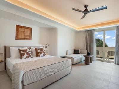 Modern hotel room with large bed, sofa, ceiling fan, and balcony overlooking outdoors.