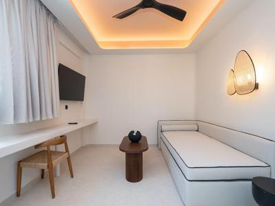 Bright, modern hotel room with white sofa, desk, wooden chair, and indirect lighting.