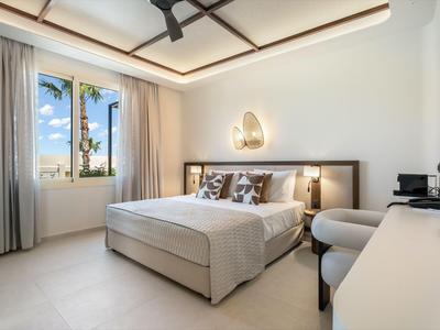 Bright hotel room with double bed, ceiling fan, and window overlooking palm trees.