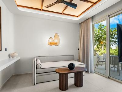 Modern hotel room with sofa, table, and balcony access overlooking greenery.