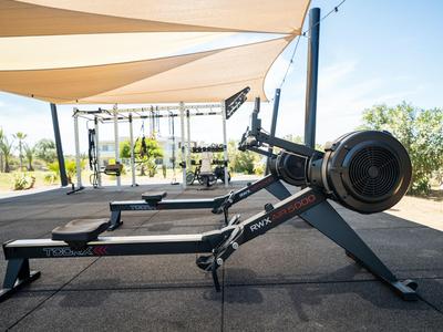 Area fitness all'aperto con vogatori sotto tende da sole accanto a palme.