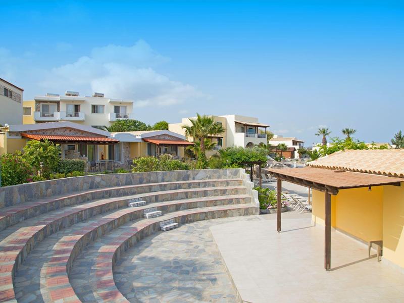 Anfiteatro al aire libre con asientos escalonados y casas en fila al fondo bajo un cielo azul.