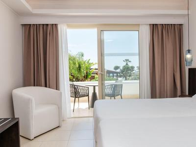 Camera d'hotel luminosa con letto matrimoniale, poltrona e balcone con vista su palme e mare.