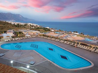 Hotelpool mit Liegestühlen, Blick auf das Meer und Berge im Sonnenuntergang.