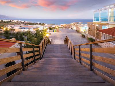 Scalinata in legno che scende verso una spiaggia con mare e edifici al tramonto.