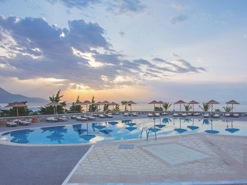 Tramonto su una piscina con lettini e ombrelloni accanto al mare con montagne sullo sfondo.
