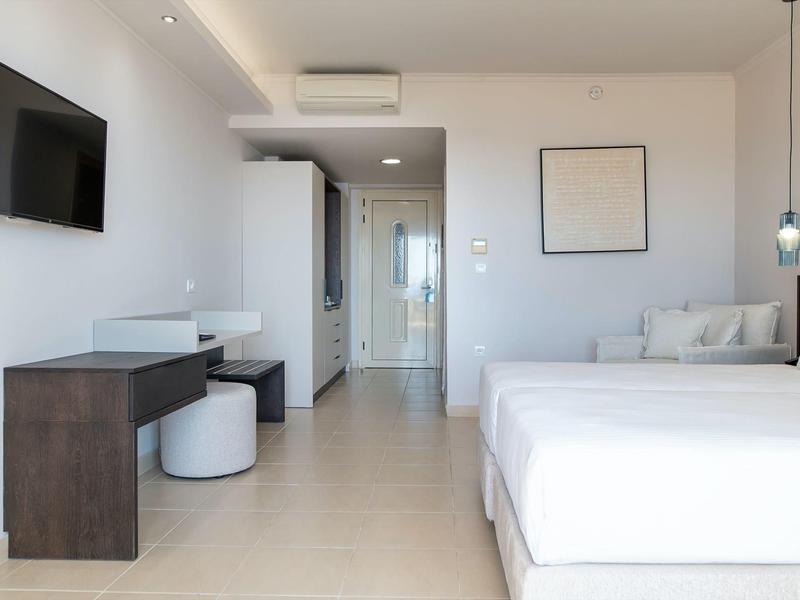 Camera d'hotel luminosa e moderna con letto matrimoniale, scrivania, divano e TV a parete.