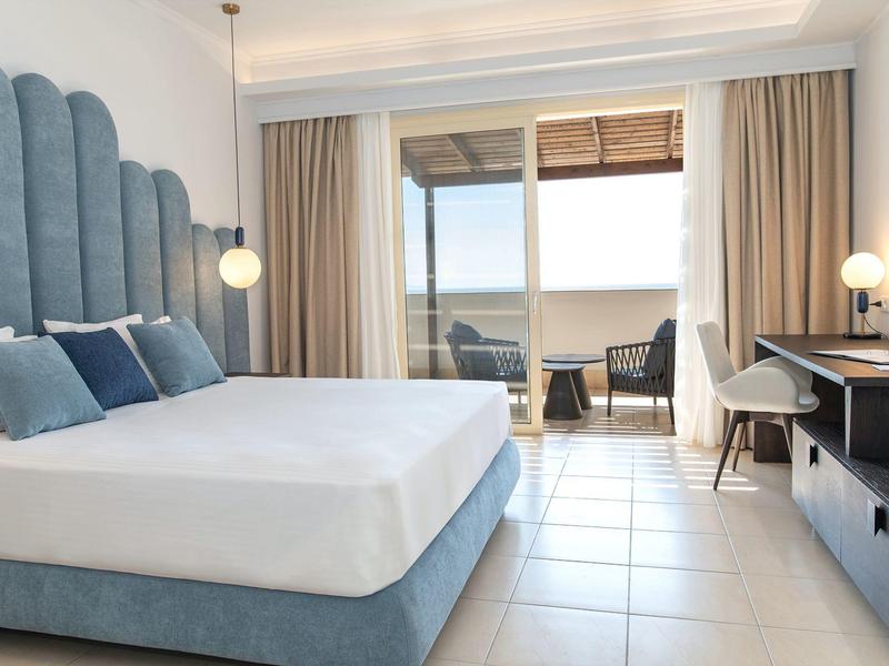 Camera d'hotel luminosa con letto grande, scrivania e balcone con area salotto.