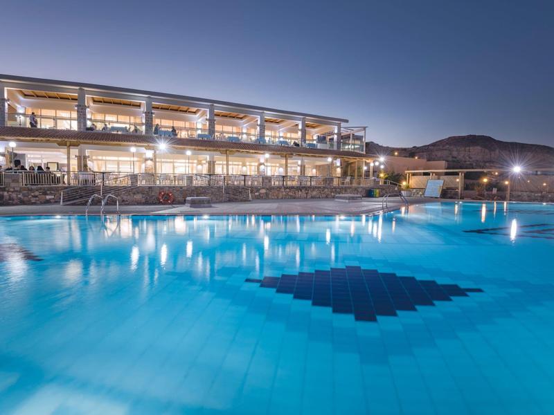 Hotel illuminato con grande piscina all'aperto al crepuscolo e montagne sullo sfondo.