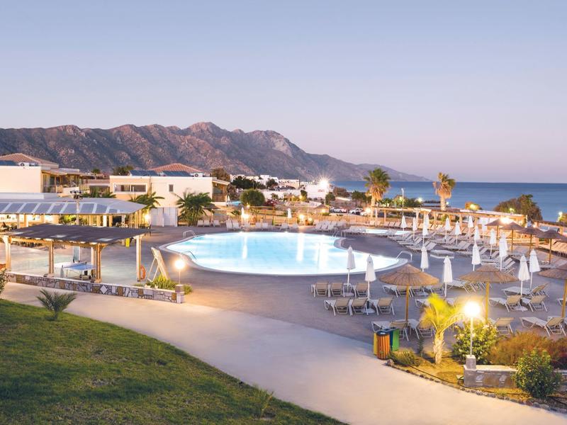 Vista serale di una piscina illuminata con lettini e montagne vicino al mare.