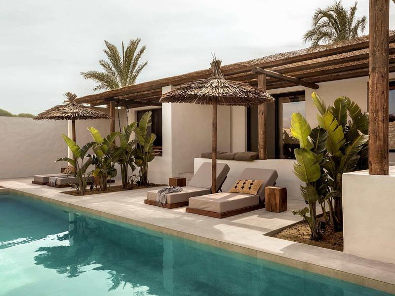 Moderner Pool mit Sonnenschirmen, Liegen und üppigen Pflanzen vor weißem Haus und Palmen.