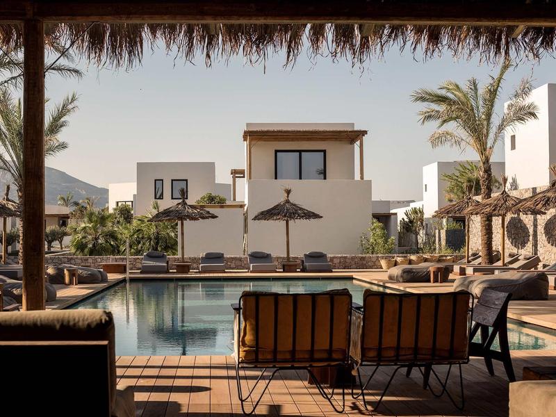 Moderner Pool mit Liegestühlen, Strohdächern und weißen Gebäuden unter klarem Himmel.