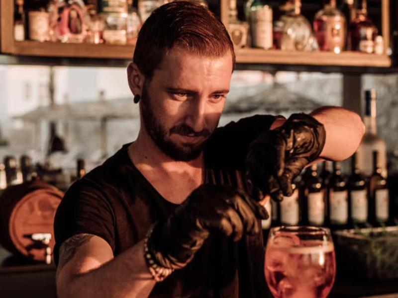 Mann mit schwarzen Handschuhen gießt Drink in großes Glas an gemütlicher Bar.