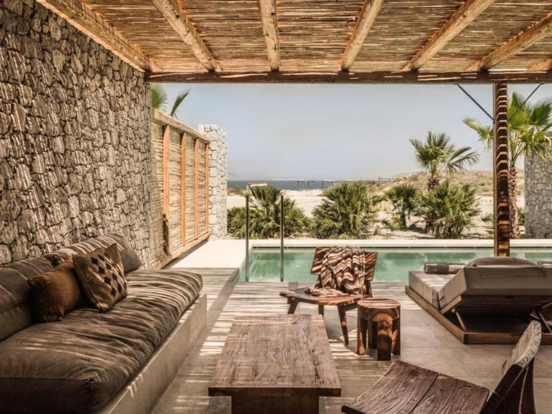 Offener Lounge-Bereich mit Steinwänden, Holzmöbeln, Pool und Blick aufs Meer bei bewölktem Himmel.