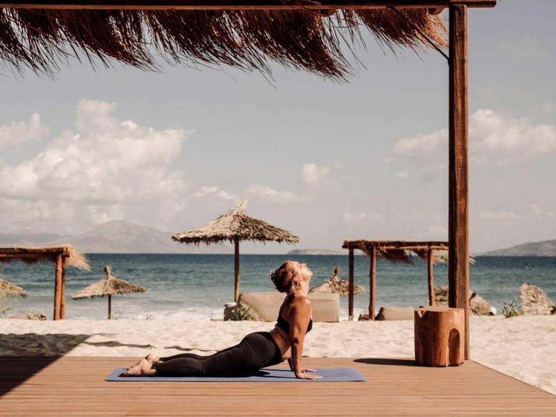 Frau praktiziert Yoga auf Holzboden am Strand mit Blick auf Meer und Inseln im Hintergrund.