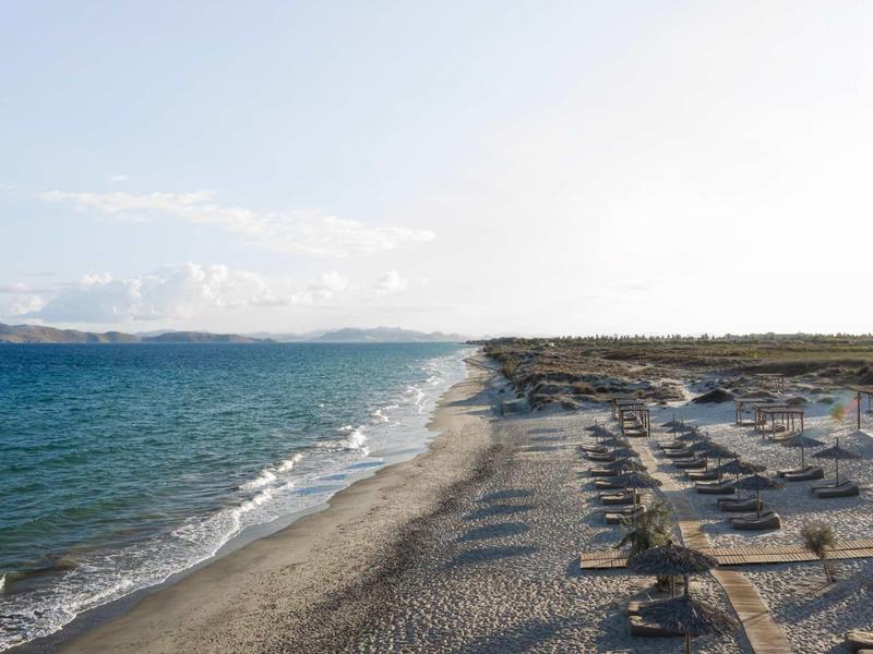 Langer Sandstrand mit vielen Sonnenschirmen und Liegen neben ruhigem blauem Meer und klarem Himmel.