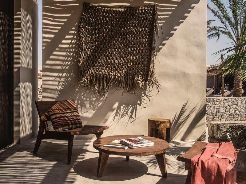 Sonnendurchflutete Terrasse mit Holzmöbeln, Tisch, dekorativen Kissen und Textil am Stuhl.