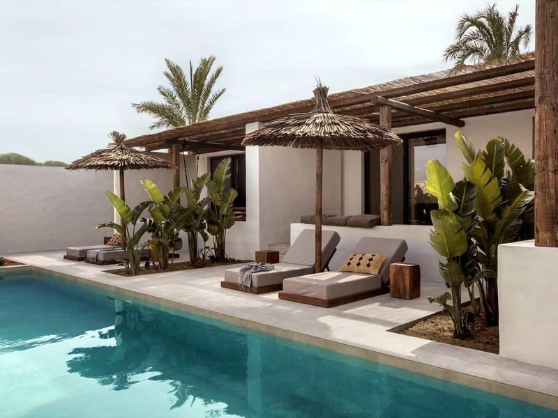 Moderner Außenbereich mit blauem Pool, Holzliegen, Palmen und weißer Mauer in ruhiger Atmosphäre.