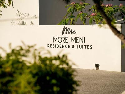 Weiße Wand mit dem Schriftzug „More Meni Residence & Suites“ unter grünen Pflanzen an sonnigem Tag.