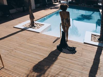 Frau mit Sonnenhut steht auf Holzterrasse neben klarem Pool in sonnigem Hotelgarten.