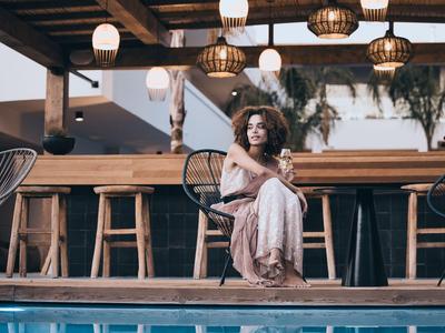 Frau in elegantem Kleid sitzt am Rand eines Pools vor einer Holzbar mit hängenden Lampen.
