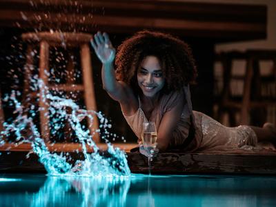 Frau entspannt sich am Poolrand mit einem Getränk und spritzt Wasser spielerisch ins Wasser.