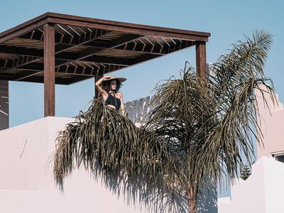 Frau mit Hut auf der Terrasse eines modernen Gebäudes, umgeben von Palmen unter blauem Himmel.