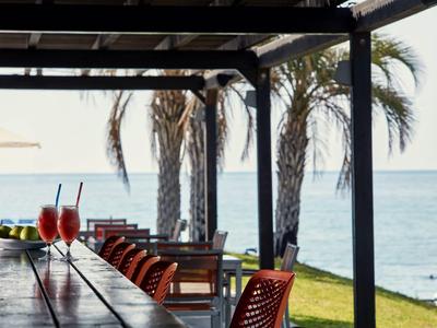 Terrazza con tavoli di legno, sedie rosse e vista sul mare con palme sullo sfondo.