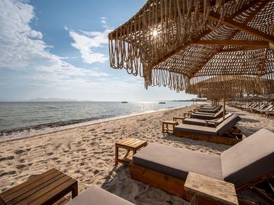 Reihe von Sonnenliegen mit Strohdach am Sandstrand vor ruhigem Meer unter blauem Himmel.