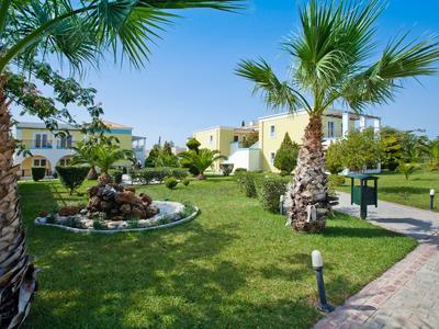 Groene tuin met palmbomen, pad en witte gebouwen onder een blauwe lucht.