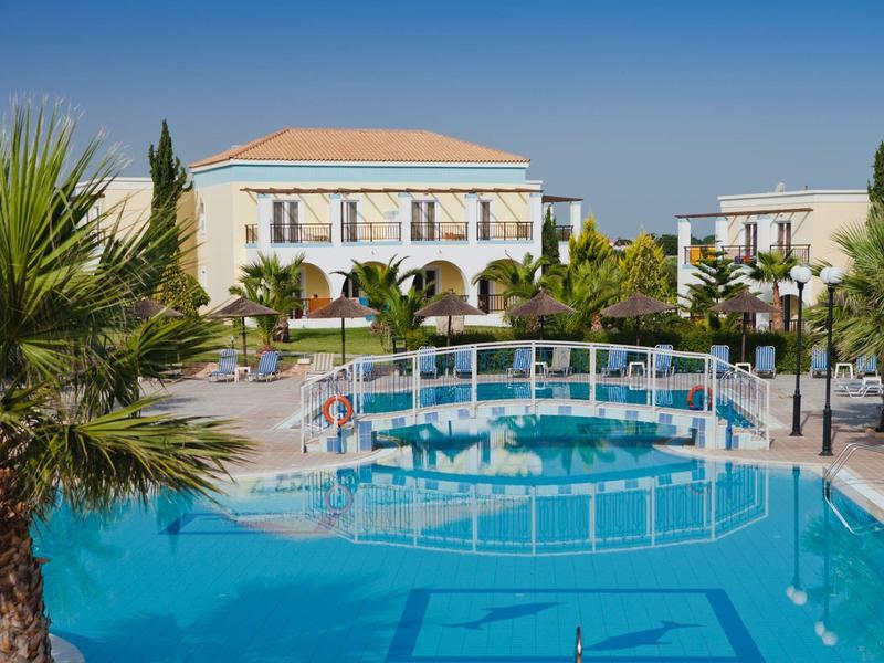 Hotelanlage mit Pool, Brücke und mediterranen Gebäuden unter klarem blauem Himmel.