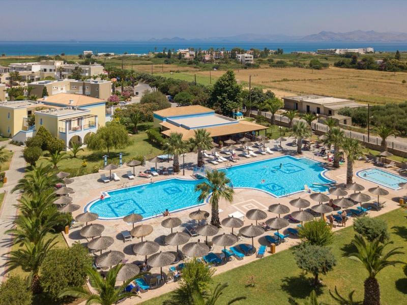 Großer Poolbereich mit Sonnenschirmen, Liegestühlen und umliegender Hotelanlage nahe dem Meer.