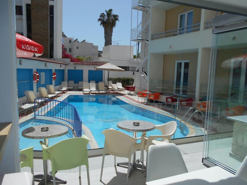 Moderner Außenpool mit weißen Stühlen, Tischen und Sonnenschirmen zwischen zwei Gebäuden.