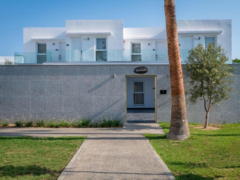 Modern white hotel building with glass railing, palm trees, and well-maintained lawn.