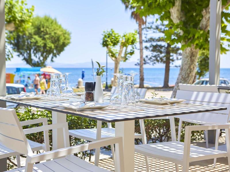 Terras met witte stoelen en tafels met uitzicht op zee onder bomen.