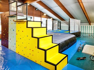 Indoor-Trampolin mit gelbem Kletterblock und Treppe, blauer Matte, Holzbalken und großen Fenstern.