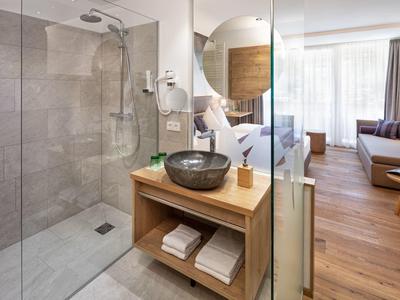 Modernes Badezimmer mit Glasdusche, schwarzem Waschbecken, Holzregal und Blick ins Wohnzimmer.