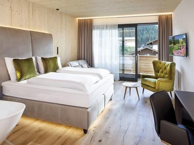 Modernes Hotelzimmer mit großem Bett, grünem Sessel, Balkon und hellem Holzboden.