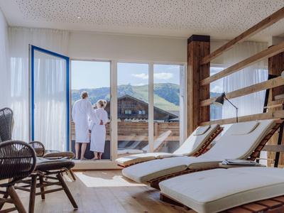 Helle Wellnesslounge mit Holzliegen, Wandleiter und Paar in Bademänteln vor Terrassentür.