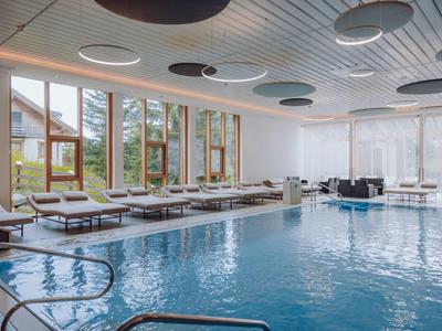 Innenraum eines Schwimmbads mit großem Becken, Holzrahmenfenstern und Liegestühlen am Rand.