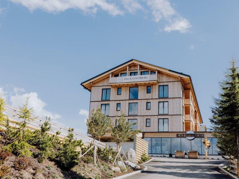 Mehrstöckiges Holzhaus auf Hügel mit Bäumen, bewölktem Himmel und Einfahrt im Vordergrund.