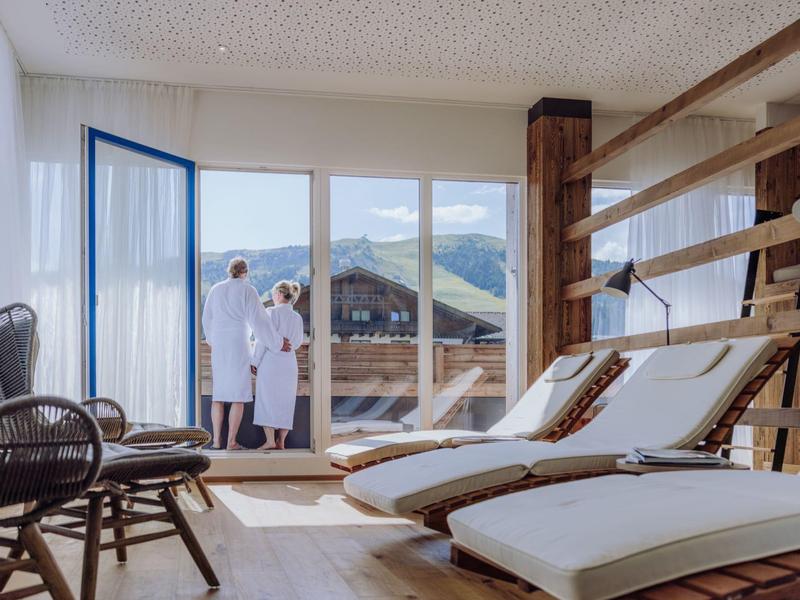 Helle Wellnesslounge mit Holzliegen, Wandleiter und Paar in Bademänteln vor Terrassentür.