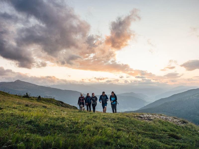 Fünf Wanderer laufen auf einem grasbewachsenen Hügel vor einem bergigen Sonnenuntergang.
