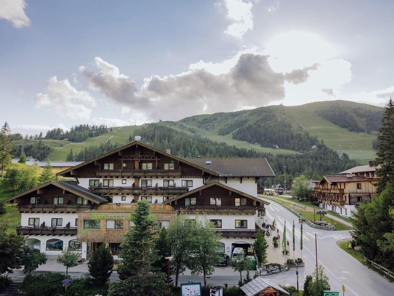 Alpine lodge with balconies surrounded by pine trees and mountain landscape under a cloudy sky.