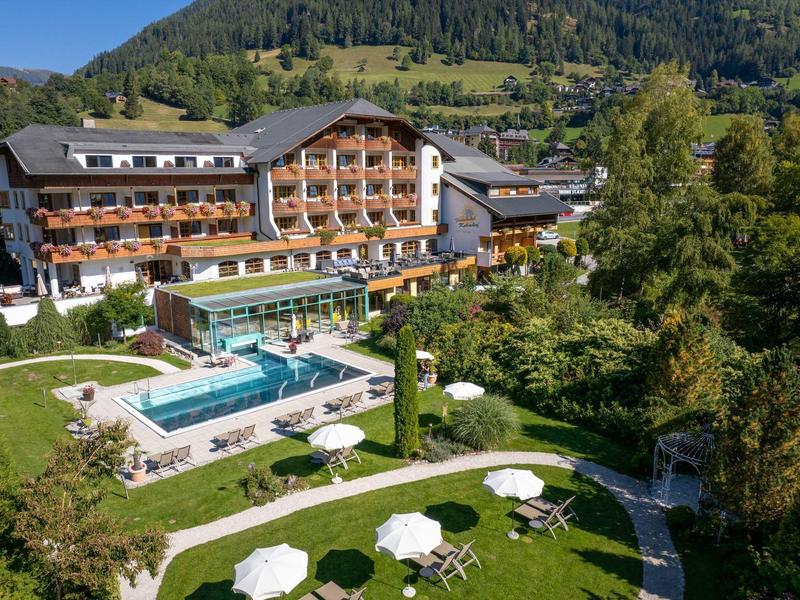 Großes Hotel mit Holzfassade, Pool und Sonnenschirmen, umgeben von grünen Bäumen und Bergen.