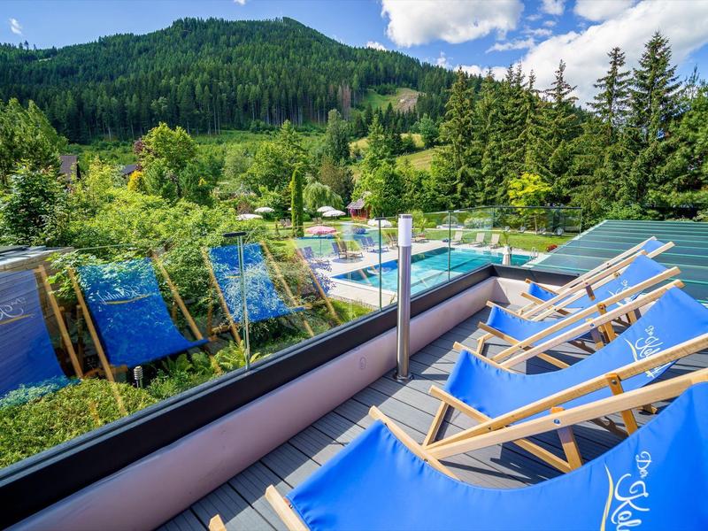 Terrasse mit blauen Liegestühlen, Glasgeländer, umgeben von grünen Bäumen und Bergen im Hintergrund.