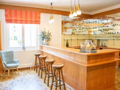 Heller Raum mit Holztresen, fünf Hockern und einem blauen Sessel neben Fenster mit oranger Gardine.